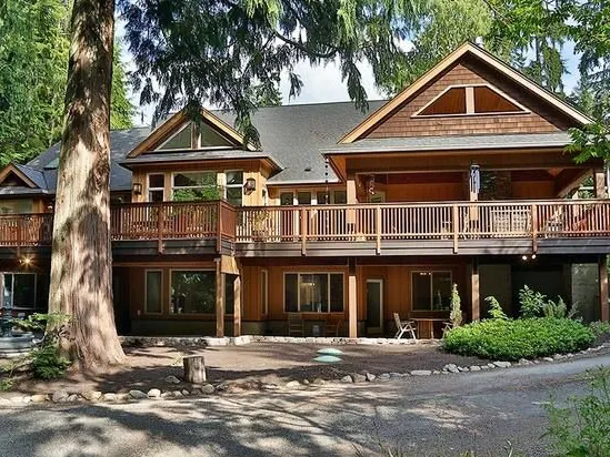Front of a custom home with cedar shingle gables and a long deck under tall evergreens