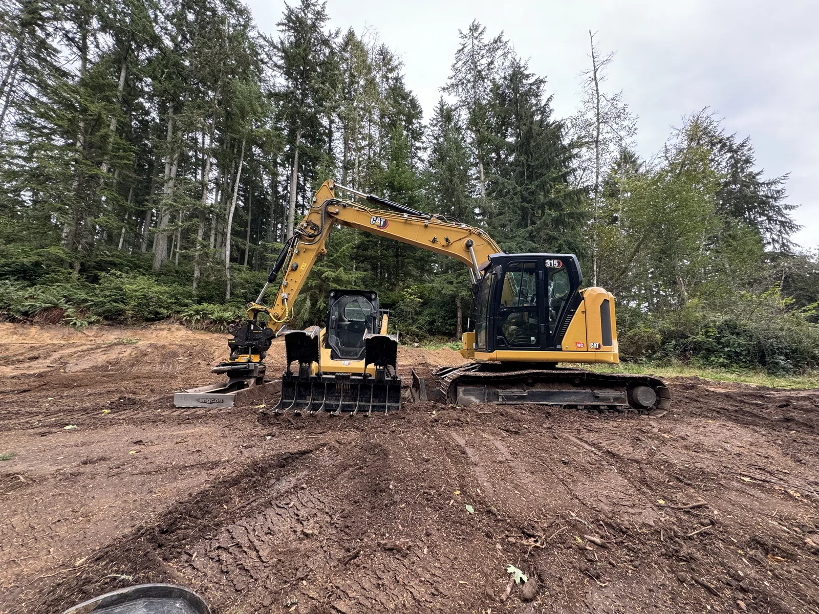 Excavator grading a cleared building site with evergreens behind