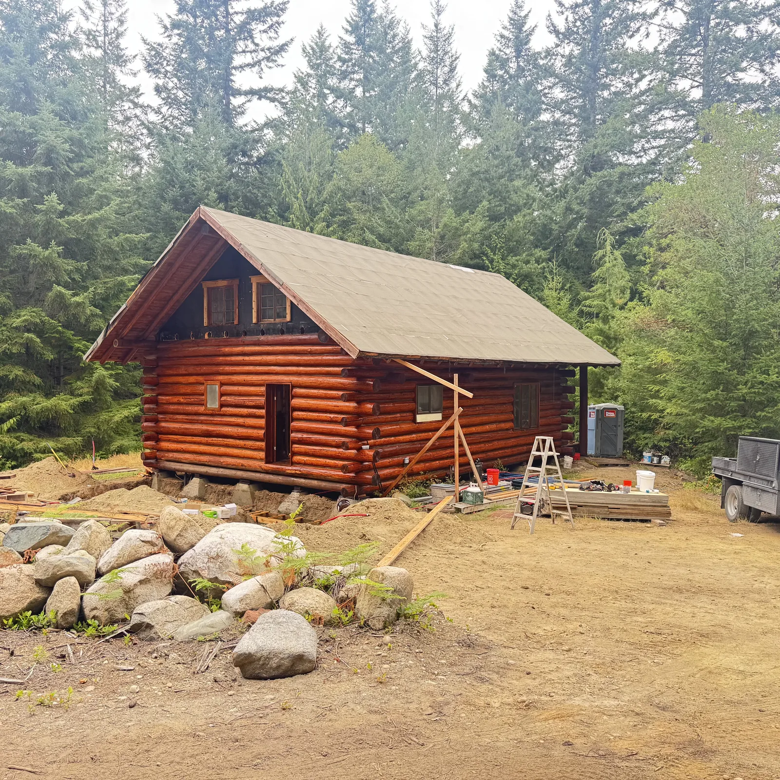 Log cabin under construction with boulders staged in front on a cleared site
